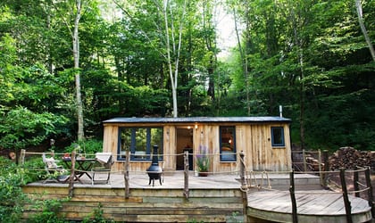 A wooden cabin with an outside deck area with a table and chairs in the middle of a forest