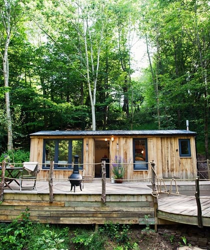 A wooden cabin with an outside deck area with a table and chairs in the middle of a forest