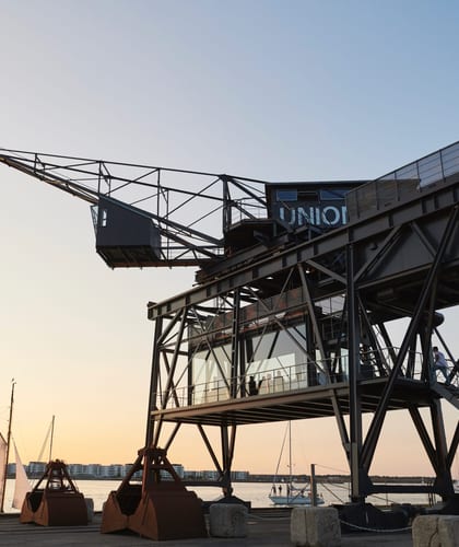 A building on stilts with a crane at the top next to a body of water with three sailing boats