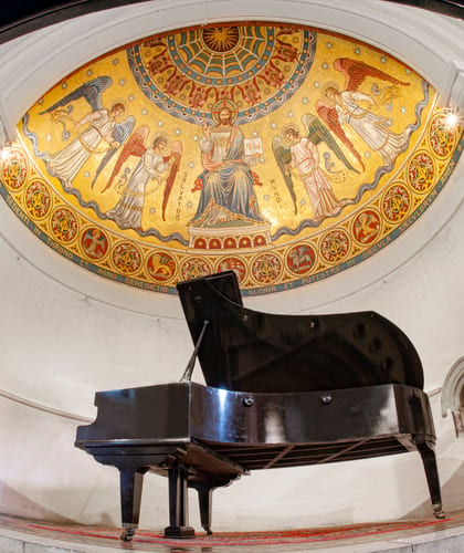 Black grand piano forte inside a religious chapel with ornate red and gold religious paintings on the domed ceiling